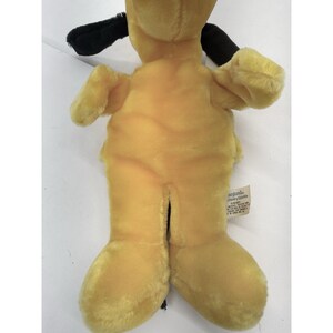 Puede incluir: Un juguete de peluche con forma de perro con pelaje amarillo y orejas negras. El juguete tiene una etiqueta adjunta en la parte trasera. El cuerpo del perro es de color amarillo liso y sus orejas son negras.