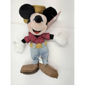 K&ouml;nnte beinhalten: Ein Mickey Mouse Pl&uuml;schtier mit einem weinroten Hemd, einer blauen Jeans und einem beigefarbenen Cowboyhut. Das Pl&uuml;schtier hat schwarze Ohren, ein wei&szlig;es Gesicht und braune Schuhe. Der G&uuml;rtel ist gelb mit einer goldenen Schnalle. Ein rotes Etikett ist angebracht.