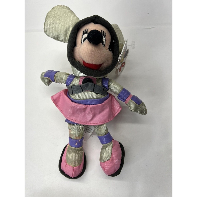 May include: A plush toy of Minnie Mouse dressed in a silver and purple space suit with a pink skirt. She has a black helmet with a white circle on the front.