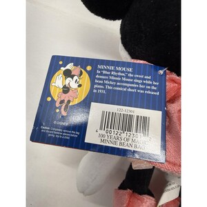 Puede incluir: Un peluche de Minnie Mouse con un vestido rosa y un lazo negro. La etiqueta presenta una ilustraci&oacute;n de Minnie Mouse y texto sobre la caricatura de 1931 "Blue Rhythm". La etiqueta tambi&eacute;n dice "100 Years of Magic Minnie Bean Bag".