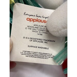 Puede incluir: Una etiqueta blanca con el texto "Everyone loves to get applause" en rojo y naranja. La etiqueta tambi&eacute;n incluye el texto "APPLAUSE IN WOODLAND HILLS, C. &copy; THE WALT DISNEY CO MADE IN CHINA 14766". La etiqueta es lavable en la superficie.