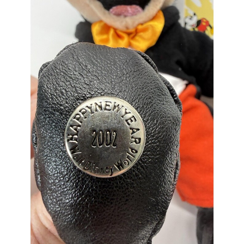 Peut inclure: Gros plan sur une peluche Mickey Mouse. Le jouet porte un smoking noir avec un n&oelig;ud papillon orange. Le bas du pied du jouet porte un bouton argent&eacute; avec l'inscription "HAPPY NEW YEAR 2002" et "Walt Disney World".