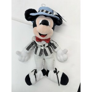 May include: A plush Mickey Mouse doll wearing a light blue hat, a red bow tie, and a black and white striped jacket. The doll has white gloves and pants, and black and white striped shoes. The doll is a collectible item.