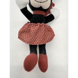 May include: A plush toy mouse with black fur, white gloves, and red shoes. The mouse wears a red polka-dot skirt and a matching scarf. The toy has a smiling face and is a classic character.