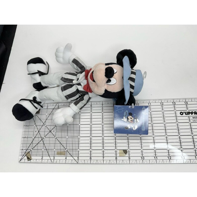 May include: A Mickey Mouse plush toy in a black and white striped outfit, red bow tie, and blue hat. The plush toy is lying on a white surface next to a ruler.