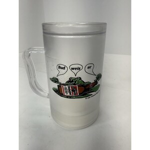 May include: Clear plastic beer mug with a handle. The mug features a cartoon frog holding a Budweiser beer bottle, with the words "Bud weis er" in speech bubbles above. The mug has a frosted appearance.
