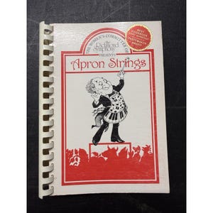 May include: A white spiral-bound cookbook titled Apron Strings, presented by The Women's Committee of the Richmond Symphony. The cover features a black and white illustration of a conductor wearing an apron with musical notes and piano keys.