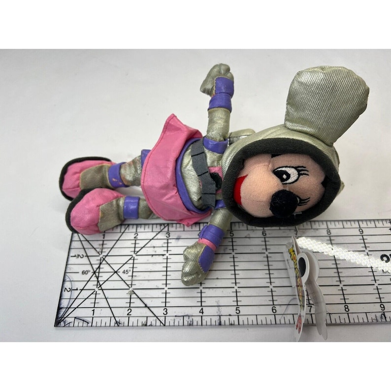 May include: A plush toy of a cartoon mouse wearing a silver and purple space suit with a pink skirt. The toy is lying on a ruler with markings in inches.