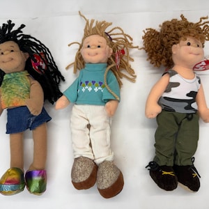 May include: Three handmade rag dolls with smiling faces. The dolls have different hairstyles, clothing, and shoes. One doll has black yarn hair, a denim skirt, and colorful shoes. Another doll has blonde yarn hair, a blue sweater, and white pants. The third doll has brown curly hair, a camouflage tank top, and green pants.