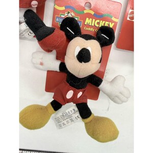 Puede incluir: Un peluche de Mickey Mouse con una etiqueta roja. El peluche tiene un cuerpo negro, pantalones cortos rojos, zapatos amarillos y guantes blancos. La etiqueta dice "MICKEY Cuddly C" y "MATT". El juguete est&aacute; hecho de fibras de poli&eacute;ster y es lavable en la superficie.