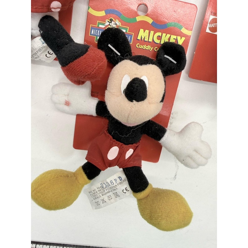 Puede incluir: Un peluche de Mickey Mouse con una etiqueta roja. El peluche tiene un cuerpo negro, pantalones cortos rojos, zapatos amarillos y guantes blancos. La etiqueta dice "MICKEY Cuddly C" y "MATT". El juguete est&aacute; hecho de fibras de poli&eacute;ster y es lavable en la superficie.