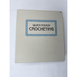 May include: A light beige book titled "Quick 'N Easy Crocheting" with a blue decorative border. The book is closed and appears to be in good condition. The text is in a simple, easy-to-read font.
