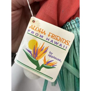 Puede incluir: Una etiqueta de "Aloha Friends From Hawaii by applause&reg;" con una ilustraci&oacute;n de flor de ave del para&iacute;so. La etiqueta est&aacute; adherida a una mu&ntilde;eca con una parte superior roja y flecos verdes.