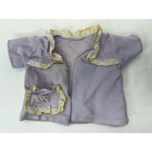May include: A lavender-colored jacket with a cream-colored lace trim. The jacket has a pocket on the left side and a decorative design on the shoulders. The jacket is made of a soft material.