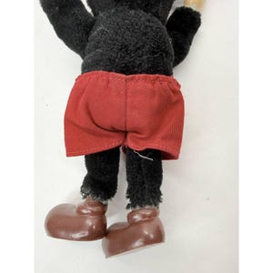 May include: A vintage plush toy featuring a black body, red shorts, and brown shoes. The toy's legs and feet are visible, showcasing its classic design. The toy's shorts are a solid red color.