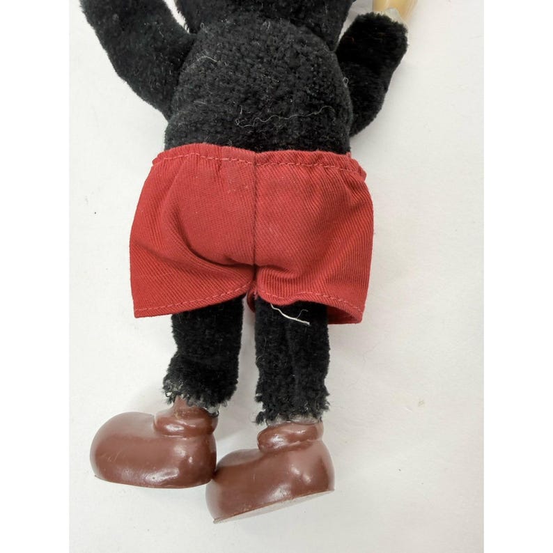May include: A vintage plush toy featuring a black body, red shorts, and brown shoes. The toy's legs and feet are visible, showcasing its classic design. The toy's shorts are a solid red color.