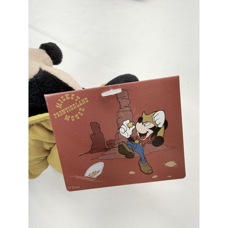K&ouml;nnte beinhalten: Ein Mickey Mouse Pl&uuml;schtier mit einem Etikett, das Mickey Mouse als Cowboy zeigt. Auf dem Etikett steht "Mickey's Frontierland Mouse" in einem kreisf&ouml;rmigen Design. Das Spielzeug hat schwarze Ohren und ein gelbes Outfit.