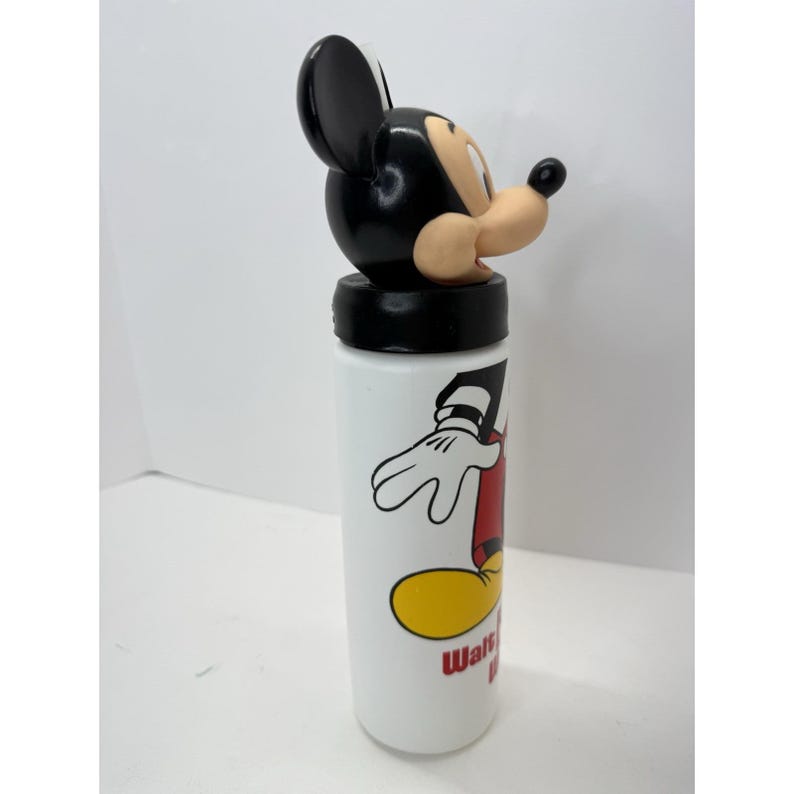 May include: White water bottle featuring a Mickey Mouse design. The bottle has a black lid and a black and tan Mickey Mouse head on top. The bottle has a cartoon graphic of Mickey Mouse and the words "Walt Disney World" in red.
