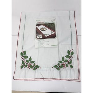 May include: A white fabric Christmas table runner featuring green holly leaves and red berries embroidered in the corners, with a thin red border. An instruction sheet titled Christmas Table Runner with a product image is placed on top.
