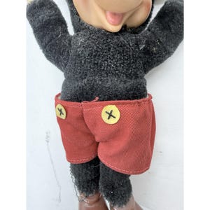 May include: A vintage Mickey Mouse plush doll. The doll has a black body and red shorts with yellow buttons. The doll's face is visible, with a smiling expression. The doll's feet are brown.