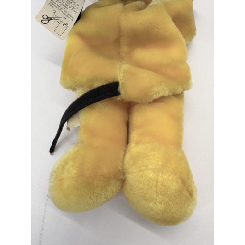 Puede incluir: Un juguete de peluche amarillo con una cola negra. El juguete tiene una etiqueta con instrucciones. El cuerpo del juguete es de un material suave y difuso. Las piernas y los pies del juguete son visibles, y descansa sobre una superficie blanca.