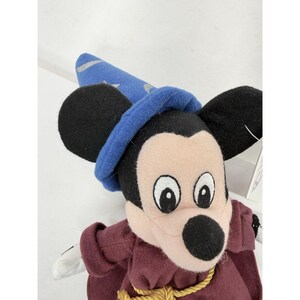 May include: A plush Mickey Mouse toy dressed as a sorcerer, wearing a blue hat with silver stars and a burgundy robe. The toy has large black ears, a white face, and a black nose. A gold rope is around the neck.