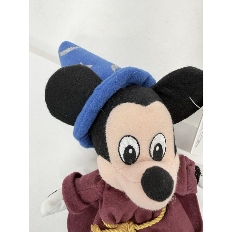 May include: A plush Mickey Mouse toy dressed as a sorcerer, wearing a blue hat with silver stars and a burgundy robe. The toy has large black ears, a white face, and a black nose. A gold rope is around the neck.