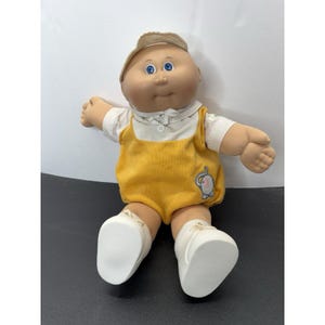 May include: A doll with a round face, blue eyes, and a tan complexion, wearing a beige cap, a white collared shirt, and yellow overalls featuring a small embroidered elephant graphic on the front, paired with white shoes with laces.