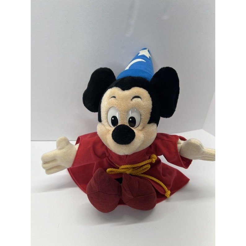 May include: A plush Mickey Mouse toy wearing a blue wizard hat and a red robe with gold trim. The toy is sitting with its legs crossed and its arms outstretched.