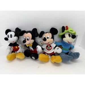 Puede incluir: Cuatro peluches de Mickey Mouse. Los peluches tienen orejas negras, caras blancas y zapatos amarillos. Un Mickey lleva un atuendo rojo y negro, otro un esmoquin, otro una camiseta blanca con un logo y el último un atuendo azul con un sombrero verde.