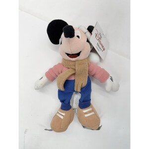 May include: A plush Mickey Mouse doll wearing a pink shirt, blue pants, and brown shoes with white stripes. The doll has a brown scarf and a black head. A tag from The Disney Store is attached.