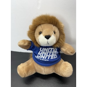 May include: A plush lion toy with tan fur and a brown mane, wearing a blue t-shirt. The shirt features the white text UNITED UNITED UNITED Van Lines printed on the front. The lion has a black nose and dark eyes, sitting in an upright position.