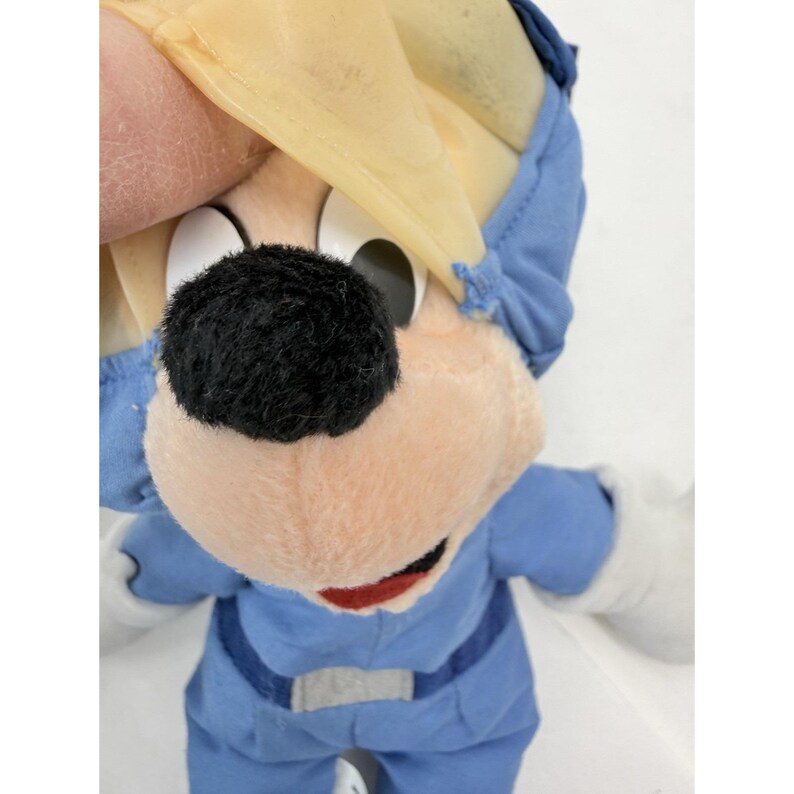 May include: A plush toy of a cartoon mouse wearing a blue jumpsuit and a yellow hat. The mouse has a black nose, white eyes, and a red mouth. The toy is made of soft, plush material.