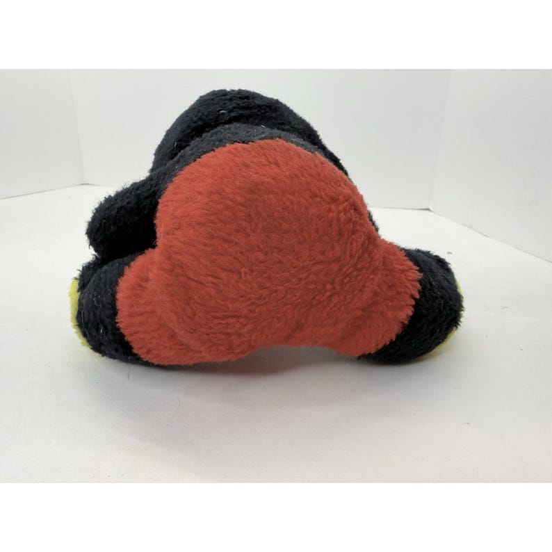 May include: A plush toy in the shape of a cartoon character, featuring a black head and limbs, and a large red body. The toy has yellow accents on the feet.