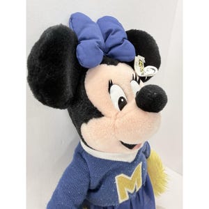 May include: A plush Minnie Mouse doll wearing a blue sweater with a yellow "M" and a matching blue bow. The doll has black ears, a pink face, and a tag attached to the ear.