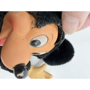 May include: Close-up of a vintage Mickey Mouse doll head. The doll has a black, fuzzy head covering, a tan face, and large white eyes with black pupils. The nose is black, and the ears are round. A tan hand is visible.