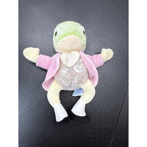 May include: A plush toy frog with a light green head, cream-colored body, and light pink jacket. The frog is wearing a paisley patterned vest and black shoes. The toy has outstretched arms and is laying on a dark surface.