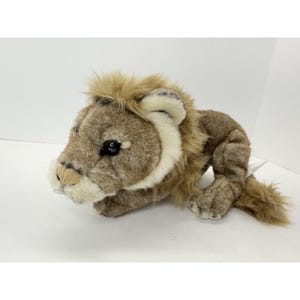 May include: A plush lion toy with a tan mane and a light brown body. The toy has a detailed face with black eyes and a beige muzzle. The lion is lying down, with its paws extended.