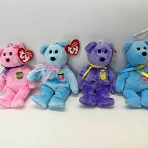 May include: Four plush Easter-themed beanie baby bears with colorful Easter egg designs. The bears are pink, blue, purple, and light blue. Each bear has a Ty tag attached to its ear.