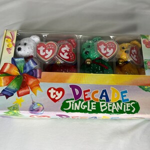 May include: A rectangular cardboard box containing four small plush bears in white, red, green, and yellow. Each bear wears a small bell and has a heart-shaped Ty tag. The box features a rainbow bow graphic and the text Ty Decade Jingle Beanies.