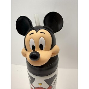 May include: A Mickey Mouse-themed tumbler with a straw. The tumbler features a black and white design with a red accent. The top of the tumbler is shaped like Mickey Mouse's head, with black ears and a tan face.