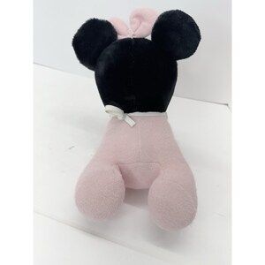 Puede incluir: Un peluche con forma de rat&oacute;n con grandes orejas negras y un lazo rosa. La cabeza del rat&oacute;n es negra, y su cuerpo y patas son de color rosa claro. Una cinta blanca est&aacute; atada alrededor del cuello.