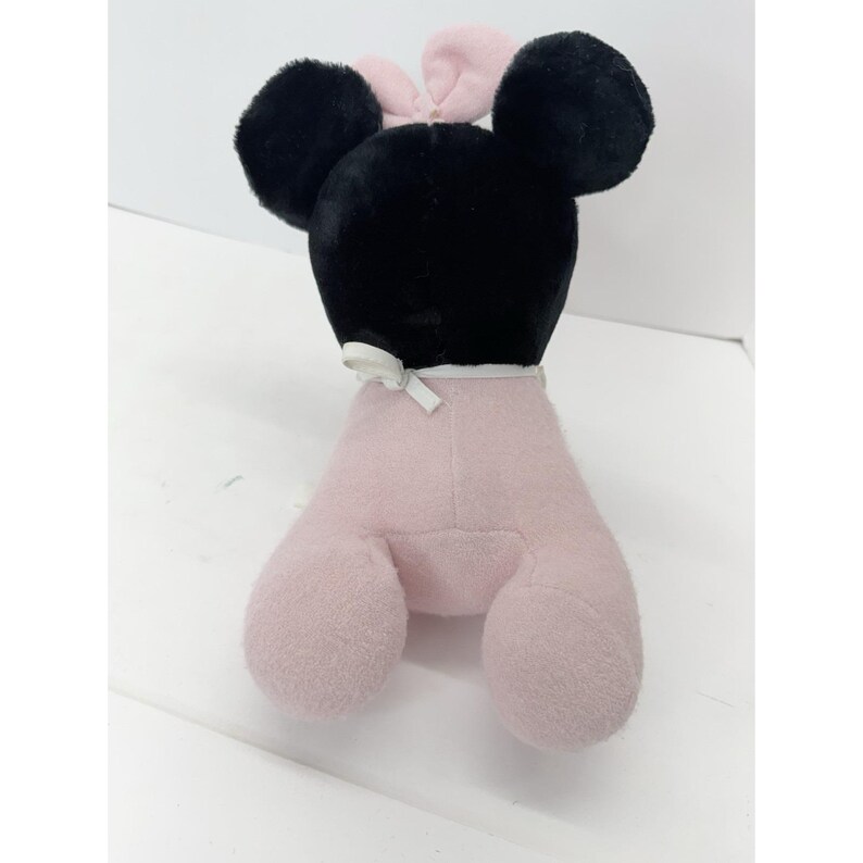 Puede incluir: Un peluche con forma de rat&oacute;n con grandes orejas negras y un lazo rosa. La cabeza del rat&oacute;n es negra, y su cuerpo y patas son de color rosa claro. Una cinta blanca est&aacute; atada alrededor del cuello.