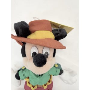 K&ouml;nnte beinhalten: Ein Pl&uuml;schtier von Mickey Mouse mit einem braunen Hut mit gelbem Band, einem gr&uuml;nen Hemd und einer roten Hose. Das Spielzeug hat schwarze Ohren und ein angebrachtes Etikett. Das Spielzeug ist ein Disney-Produkt.