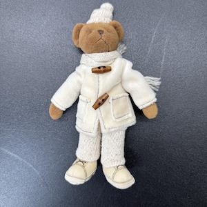 May include: A plush brown teddy bear wearing a cream-colored knit hat, a matching scarf, and a cream-colored duffle coat with two brown wooden toggle buttons. The bear is dressed in cream-colored trousers and matching cream-colored shoes.