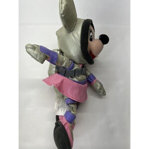 May include: A plush toy of a mouse wearing a silver spacesuit with pink and purple accents. The mouse has black eyes and a pink nose.
