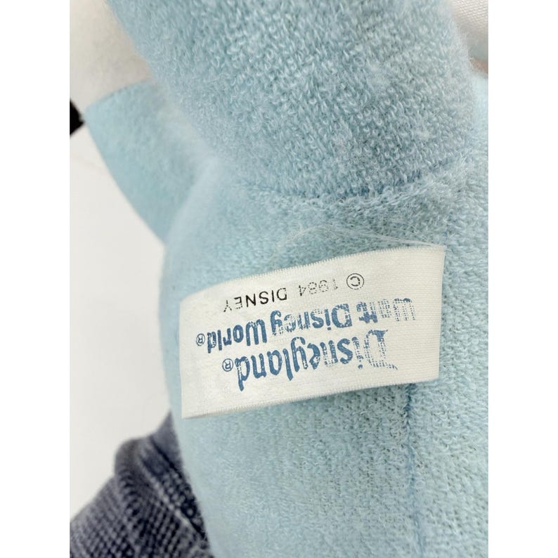 Puede incluir: Primer plano de un peluche azul claro con una etiqueta blanca. La etiqueta dice "Disneyland Walt Disney World" y "&copy; 1984 DISNEY". El peluche est&aacute; hecho de un material suave y difuso.