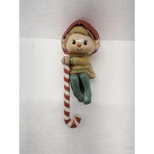 May include: A ceramic Christmas elf figurine with a red hat, holding a red and white striped candy cane. The elf has a tan tunic, green pants, and a smiling face. A festive holiday decoration.