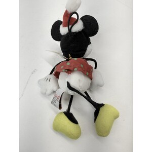May include: A plush Minnie Mouse Christmas ornament. The ornament features black ears, a red and white Santa hat, a red skirt with green polka dots, white gloves, and yellow shoes. The ornament has a black string for hanging.