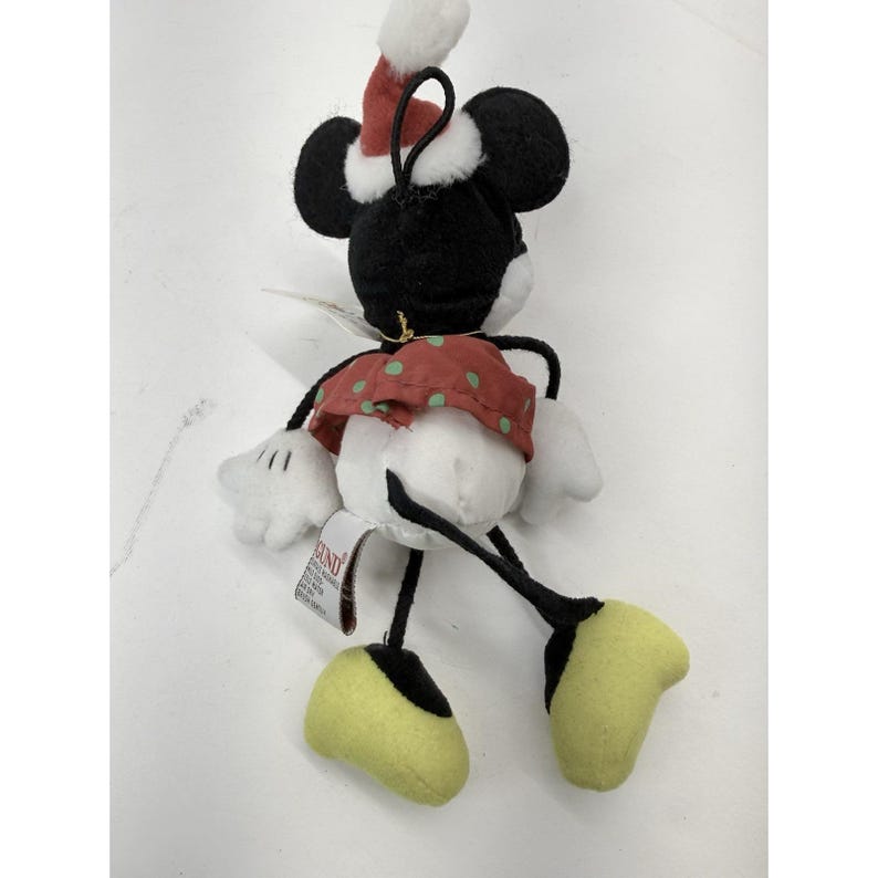May include: A plush Minnie Mouse Christmas ornament. The ornament features black ears, a red and white Santa hat, a red skirt with green polka dots, white gloves, and yellow shoes. The ornament has a black string for hanging.